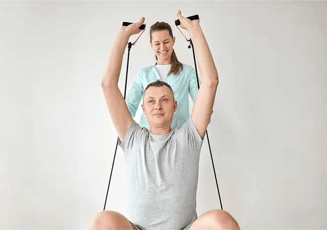 A man stretches exercise bands
        above his head while a physical therapist guides his arms
